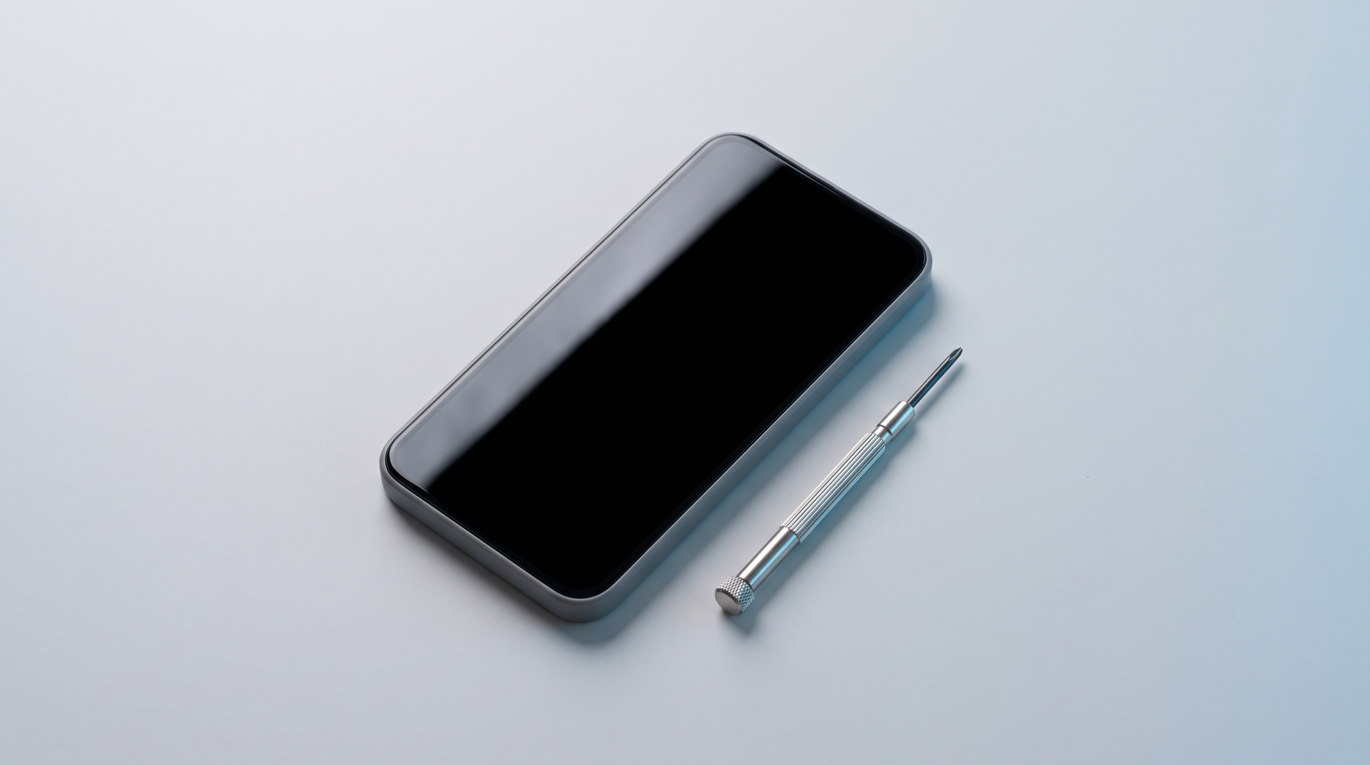 A generic, unbranded smartphone with its screen off, resting next to a small precision screwdriver on a plain pale surface.